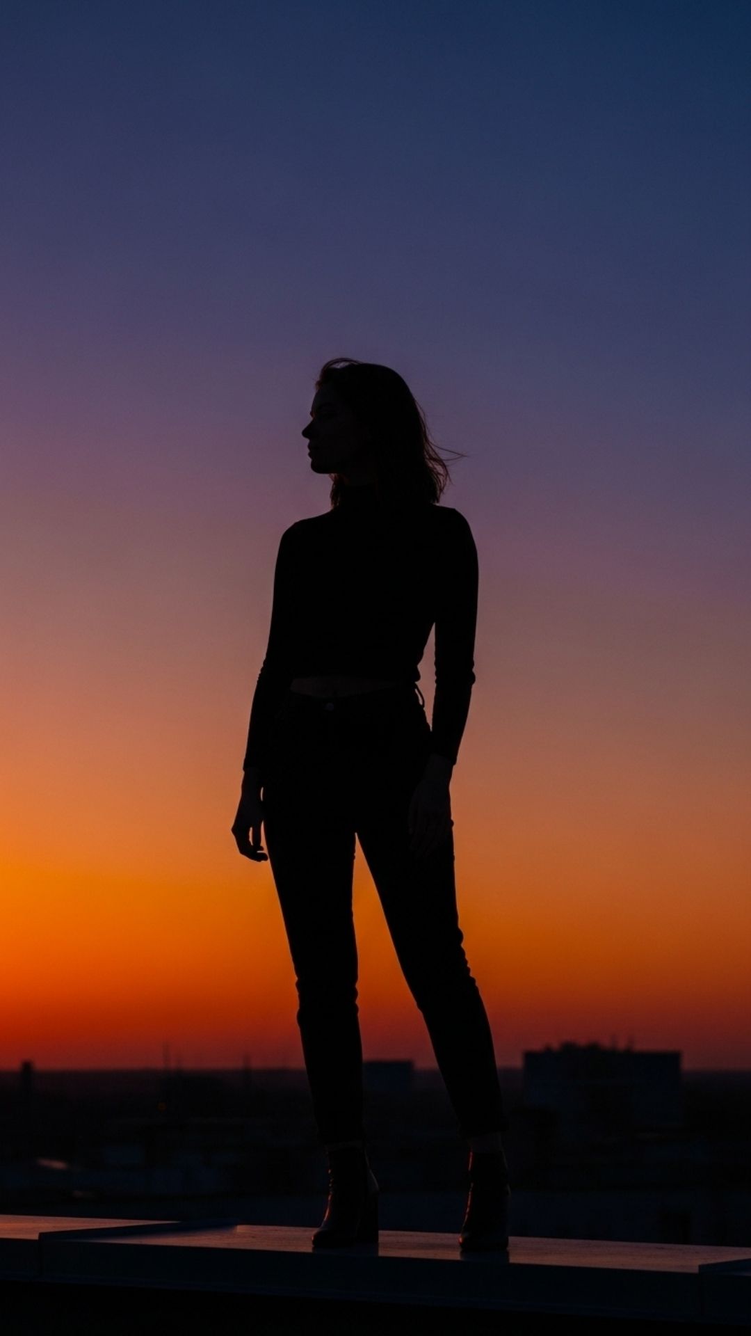 A dramatic silhouette of a young woman standing on a city rooftop against a vibrant sunset sky.