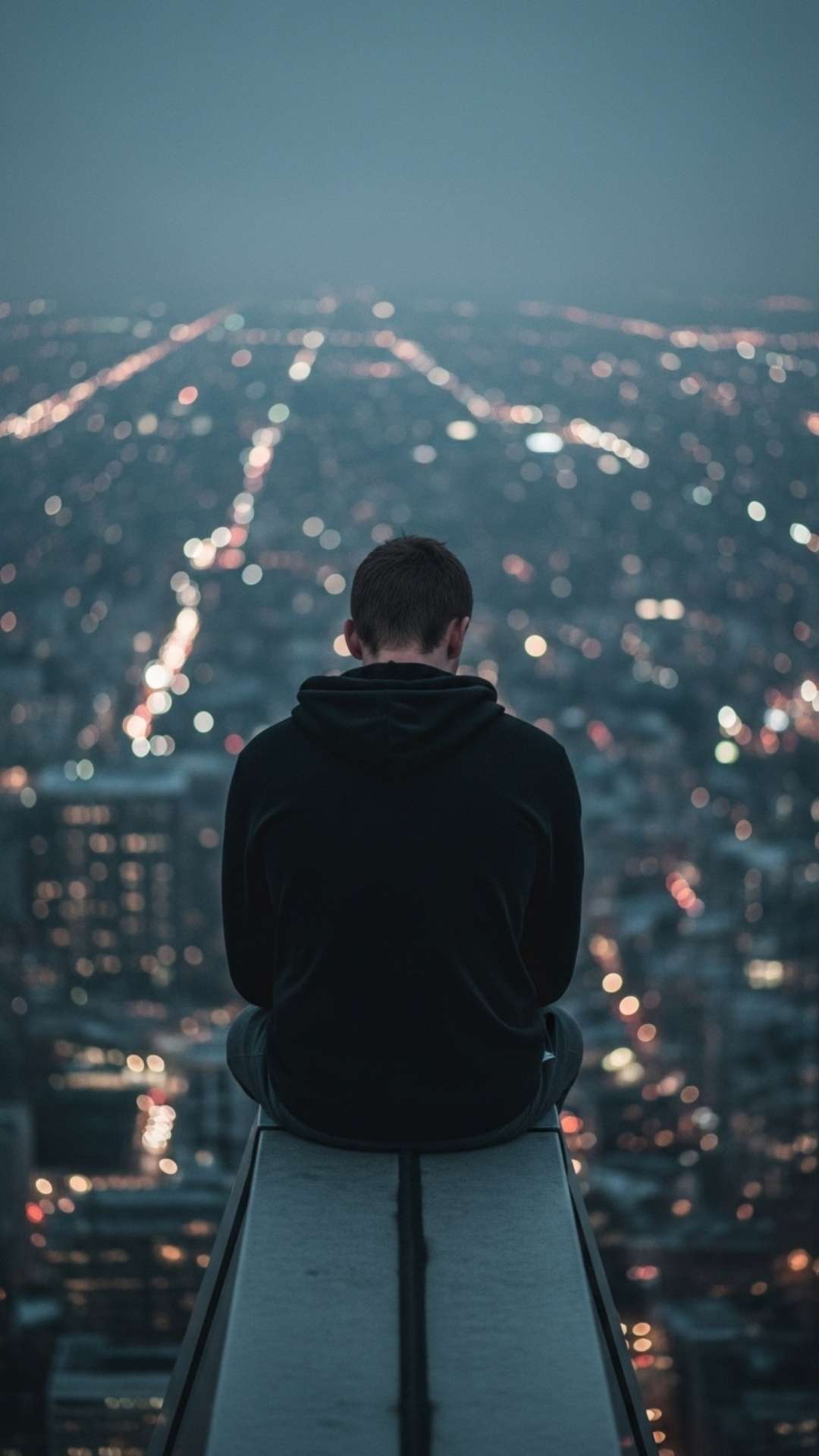A boy in a hoodie sits alone on a rooftop at night, overlooking a city skyline
