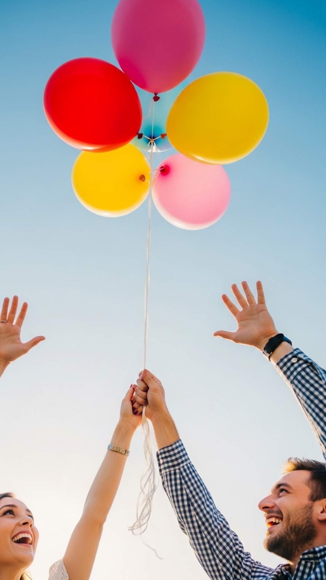 Hands of a couple holding colorful balloons against a bright blue sky, symbolizing the joyful and open expression of love.