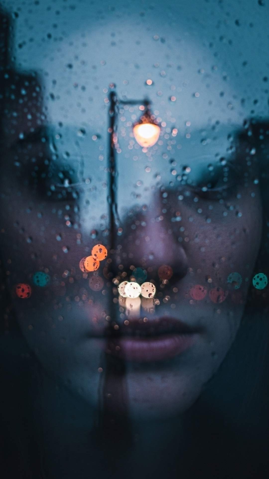 A close-up of a person's face through a rainy window, with a reflection of a streetlamp and blurred city lights
