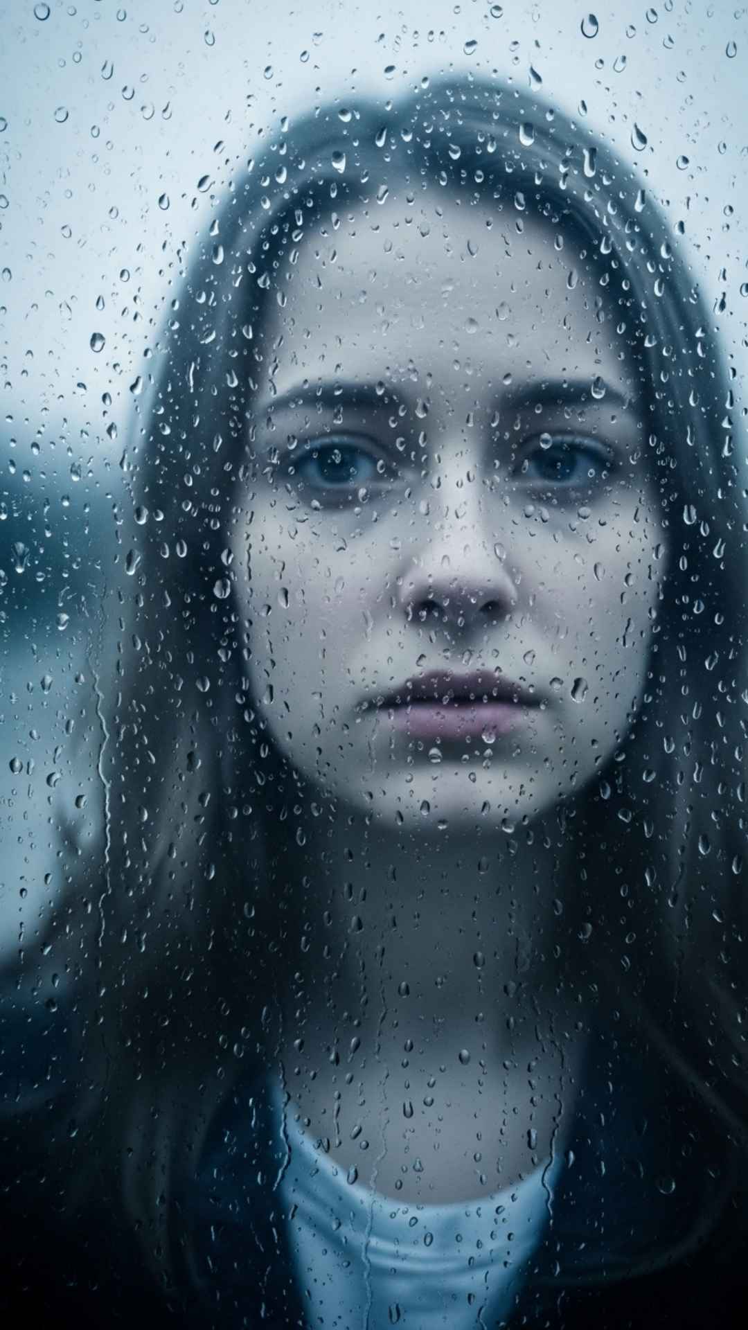 A cinematic portrait of a young woman's pensive face seen through a window covered in raindrops, in cool tones