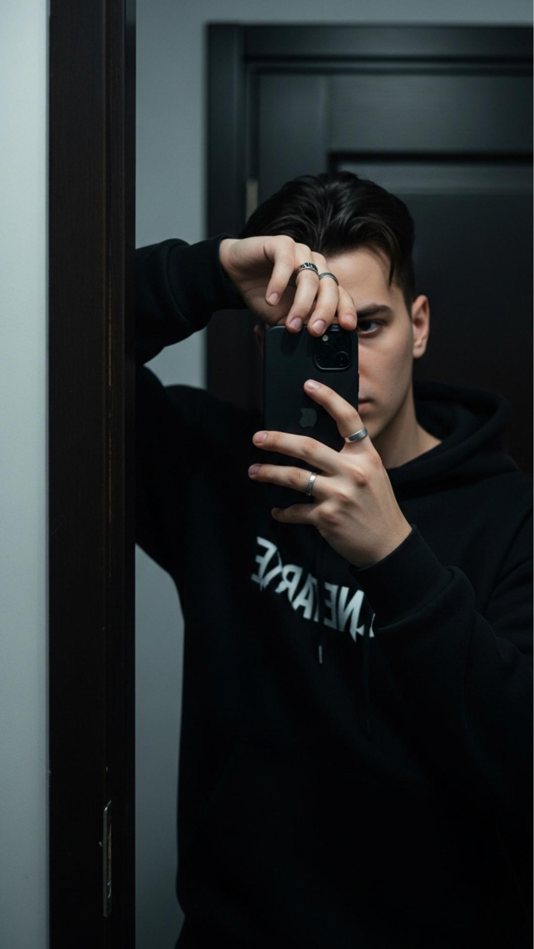 A cool mirror selfie for boys featuring a mysterious hidden face pose where the phone covers part of the face to convey attitude. The dark, moody lighting highlights the streetwear fashion, making this image ideal for users searching for anonymous and unique profile pictures for Instagram or Facebook.