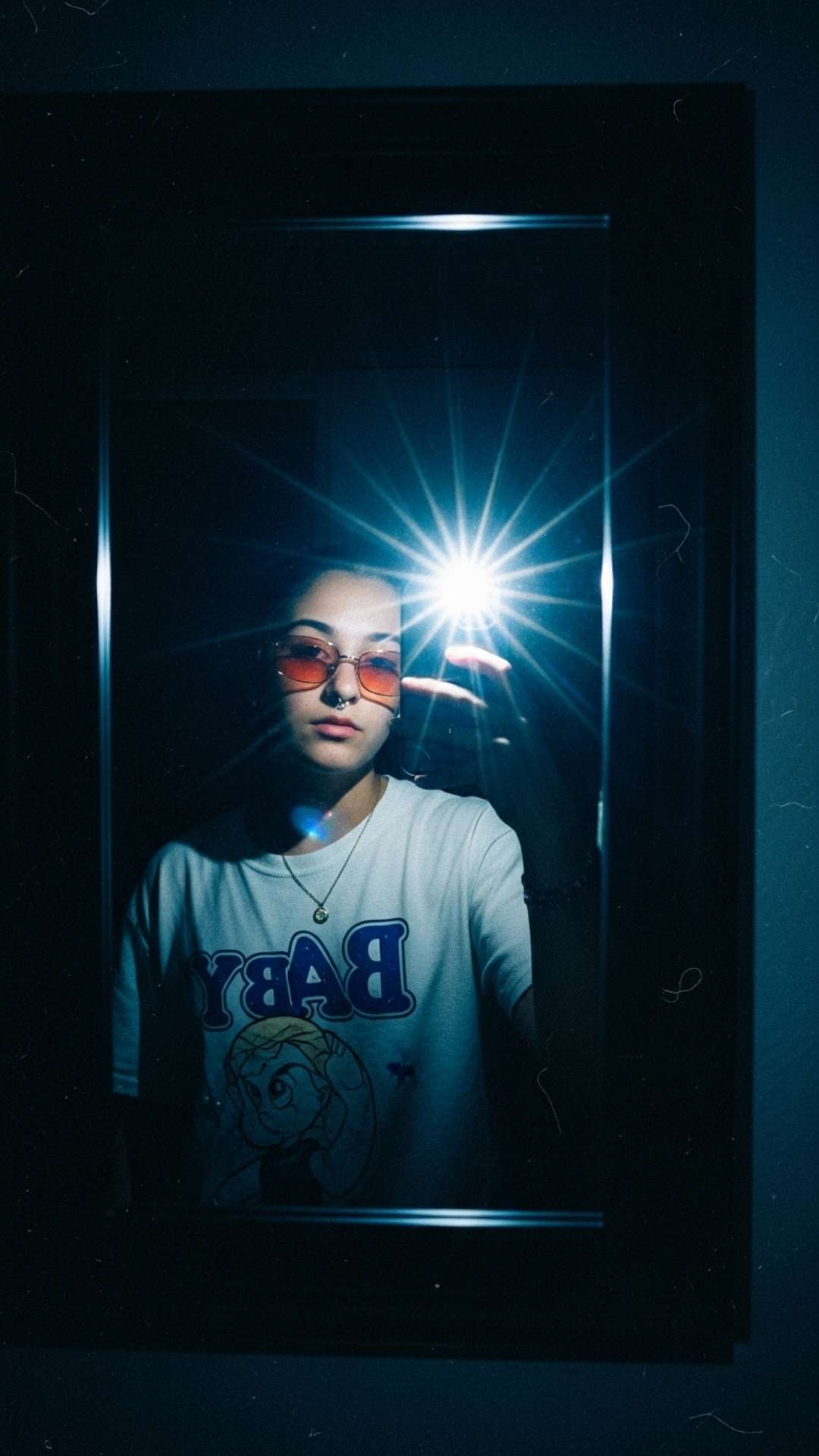 A vertical Y2K aesthetic mirror selfie taken in a dark room with a bright phone flash to create a vintage, grainy look. This "lo-fi" style is popular among Gen Z for Instagram DPs, featuring high contrast lighting and retro 2000s fashion elements like tinted glasses and digital noise